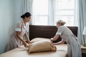 Importance of Good Housekeeping at Work Certification - Two housekeepers in uniform collaborate while making a bed in a bright, peaceful bedroom.